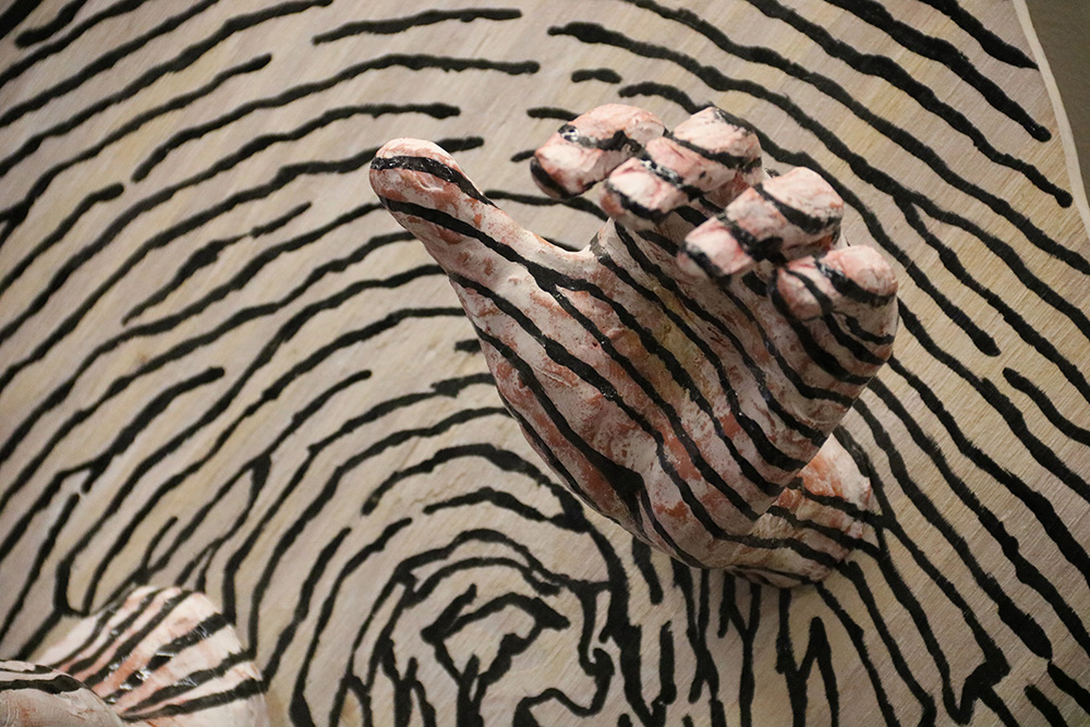 A close-up of black and white painted sculpture. There is an object resembling a human hand extruding out from the center of the sculpture. The sculpture is painted in a pattern resembling a fingerprint.