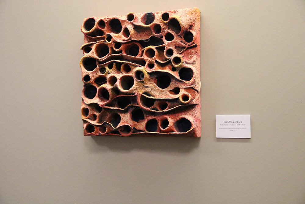A 3D orange and brown square sculpture hanging on a grey wall. The object is covered with variably sized deep holes, some of which contain smaller objects resembling human eyeballs.