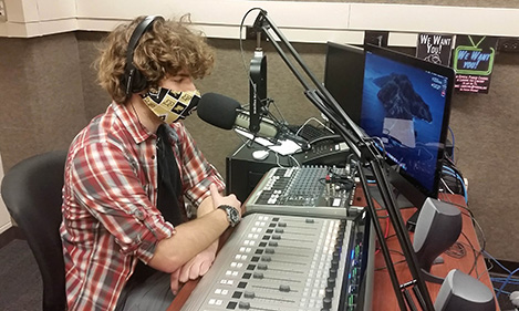 A student sitting at a desk with a mixing board and microphone recording a podcast